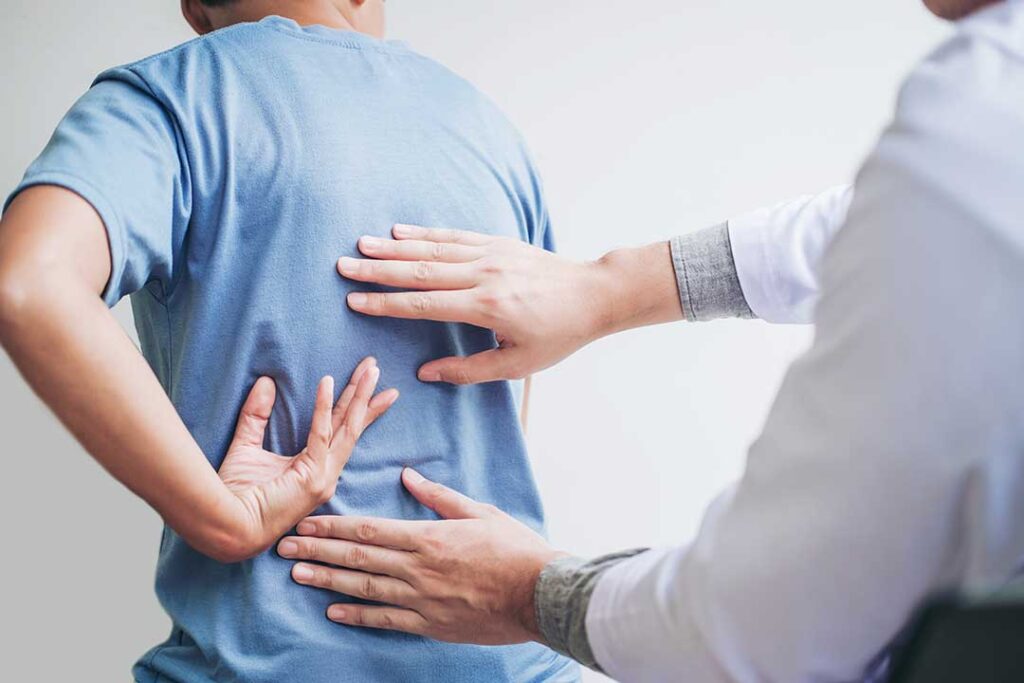 A person sits on a couch at home holding their lower back with a look of discomfort.