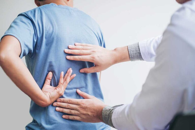A person sits on a couch at home holding their lower back with a look of discomfort.